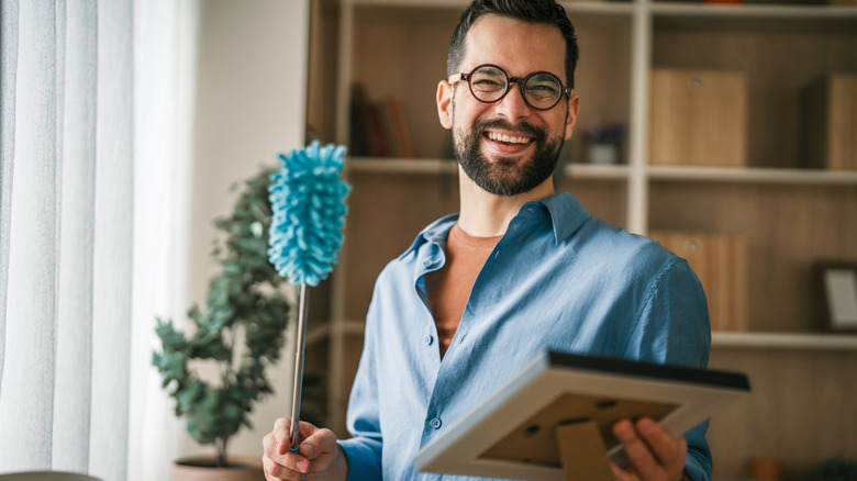 A man with glasses smiling and holding a picture frame and feather duster