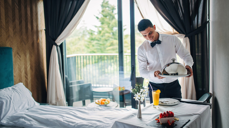 A waiter with a bowtie arranging a covered platter of food on a table in a hotel room