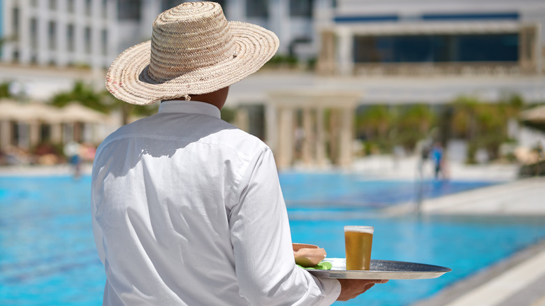 Backview of resort staff in a white shirt and straw hat delivering a glass of beer by the pool