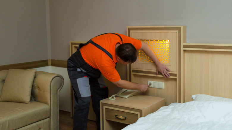 A male worker in an orange shirt fixing an electric outlet in a hotel room with a couch and bed