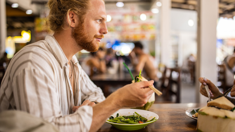 Sideview shot of a Western tourist in a striped shirt holding chopsticks over a plate of vegetables in an Asian food market