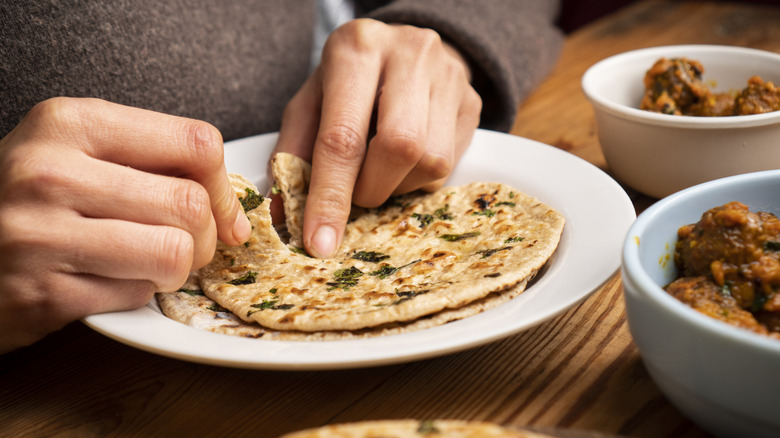 A pair of hands breaking a naan on a plate with two meat dishes on the side