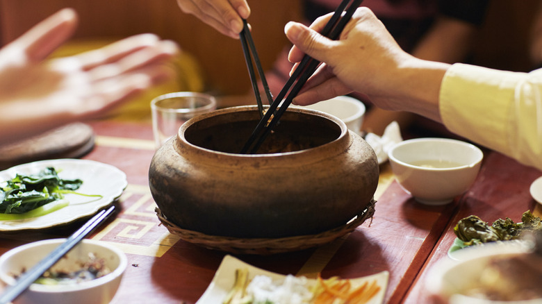 Two pairs of chopsticks dipping into a communal clay pot with small dishes and bowls in the backdrop