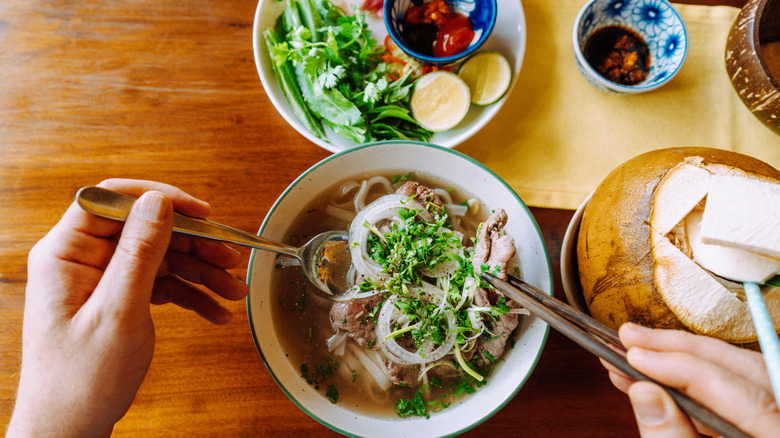 Top view of a pair of hands over a bowl of pho with condiment bowls and a coconut drink