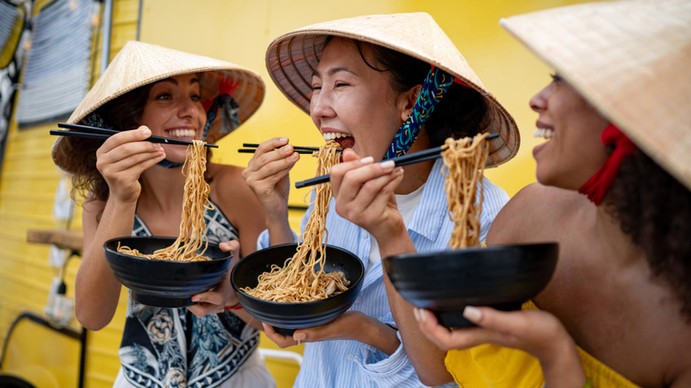 Three smiling women in Vietnamese hats eating noodles with chopsticks