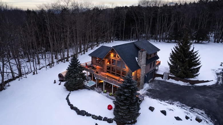 A chalet at dusk against bare winter trees. Ground is covered in snow. Three pine trees surround the house.