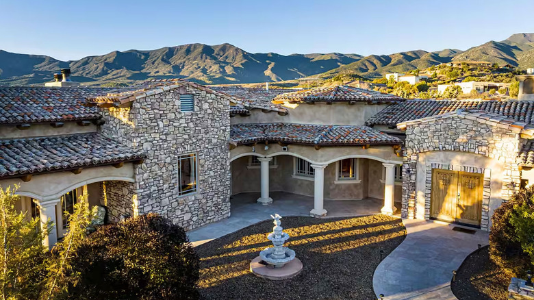 Stone-clad facades and tiled roofs of a sprawling villa against a mountain backdrop. A fountain in central courtyard.