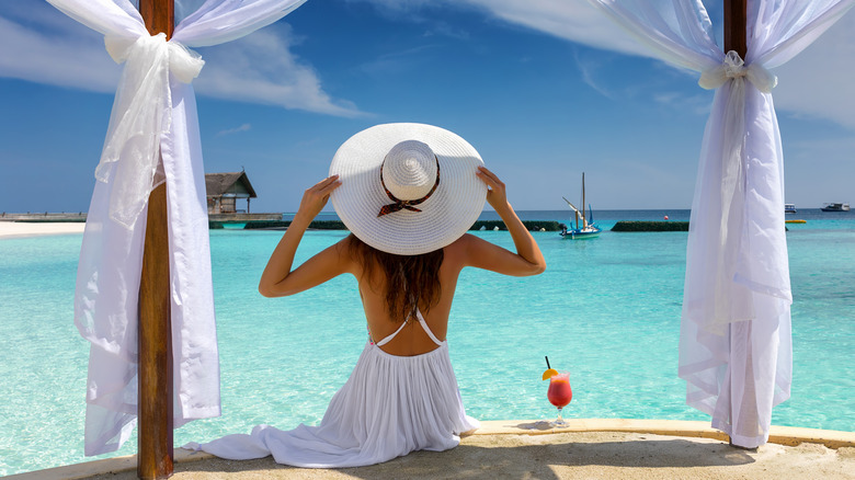 Backview of a woman in a white dress and hat, sitting on a dock facing blue seawater.