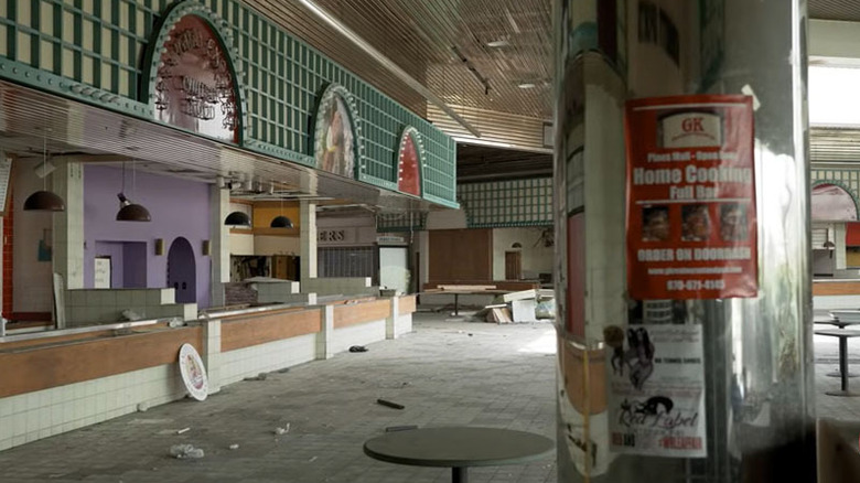 Empty food court in The Pines Mall, with a silver pillar with signs in the foreground, closed food counters along the left side, and debris-covered floors in the background
