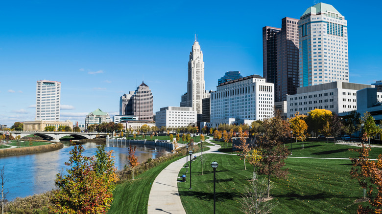 Columbus Ohio landscape showing the Scioto River to the left of a green park with autumn trees along the right foreground and skyscrapers against a blue sky in the background