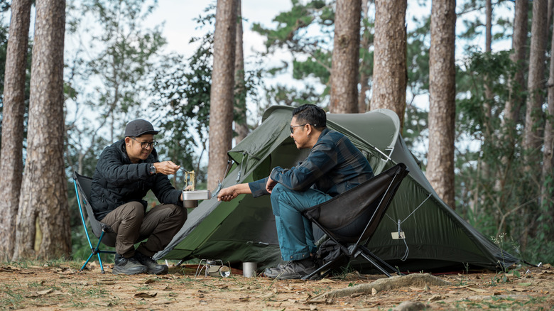 Two men share noodles at a simple campsite with a tent in the background