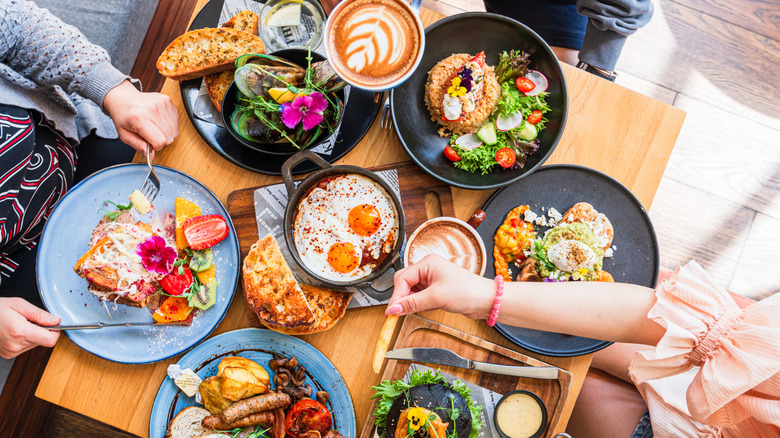 Overhead photo of a brunch spread with coffees, toasts, eggs, sausages, as hands reach for food