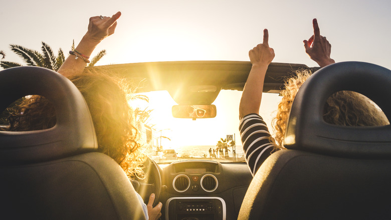 Two women singing and dancing to music in their open-roof car, as they drive along a coastal road.
