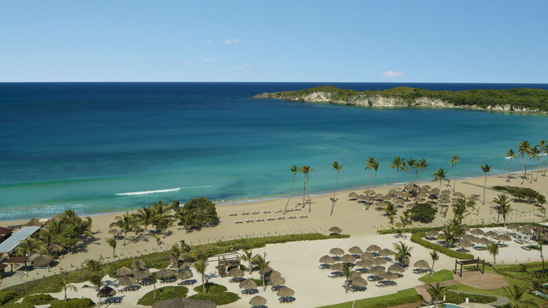 Bird's eye view of a sandy shores, dotted with plam trees, beachside huts, and hills