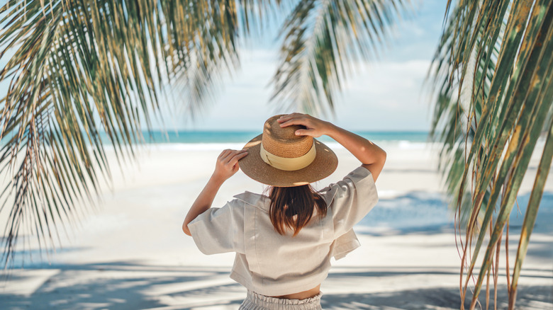 Back view of a woman holding her straw hat under palm leaves, facing a white sand beach