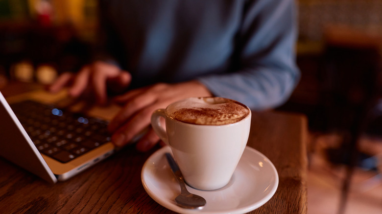 A set of hands using a laptop on a bar top with a cappuccino
