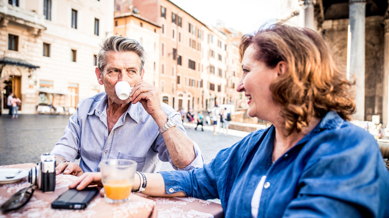 An older man and woman both in blue button downs sit at an outdoor table drinking coffee in Italy