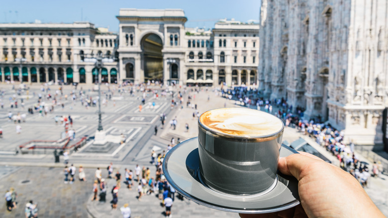 A hand holding a cappuccino with a bustling Italian plaza down below