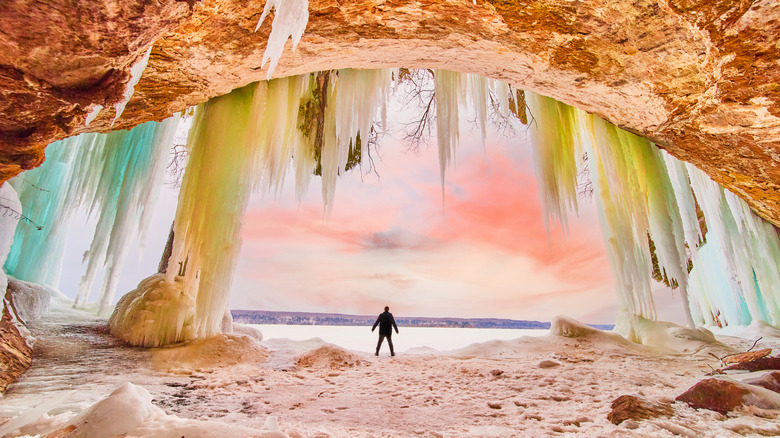 A person stands underneath dozens for colorful icicles hanging down from a cave rock face by a lake