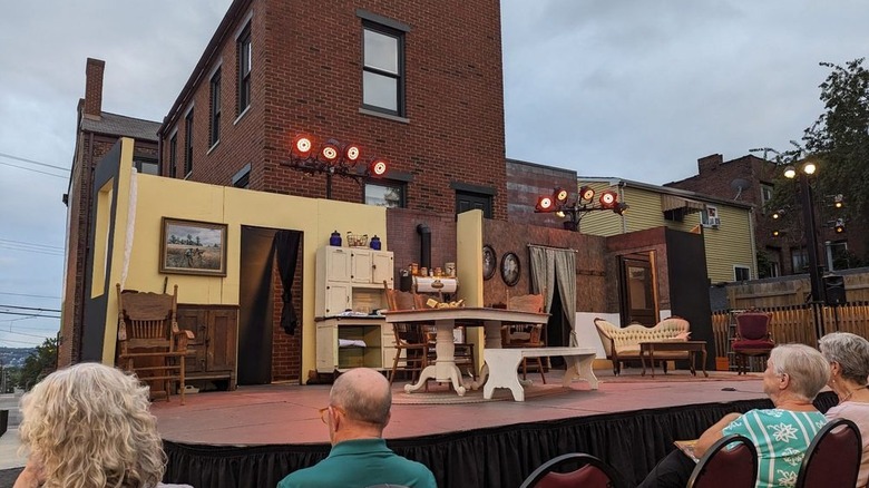 Backyard stage at the August Wilson House, with a set kitchen with yellow walls and a living room with brown walls in front of a tall brick building and audience members sitting in front of it, under a cloudy sky