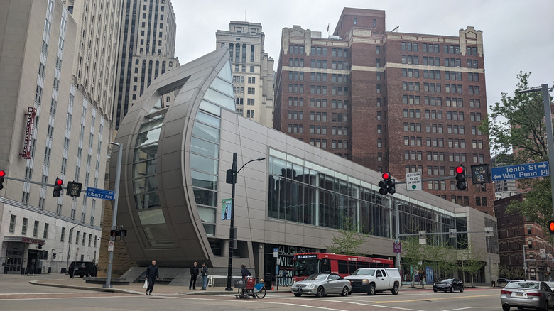 The August Wilson Center, a long gray building with large windows and a curved wall on the end closest to the camera, with a red brick building behind and other large white buildings around it, and a red bus and cars driving on the street out front