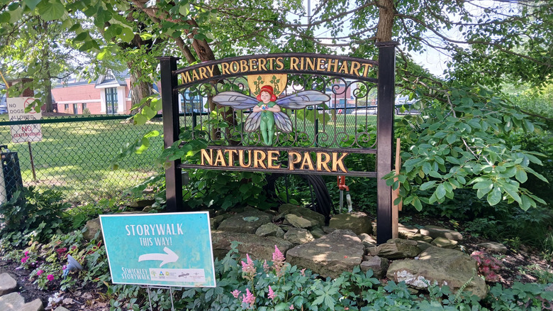 Black metal sign reading "Mary Roberts Rinehart Nature Park" in gold letters, with a fairy wearing green with blue wings, surrounded by trees and with a brick school building behind a fence in the background