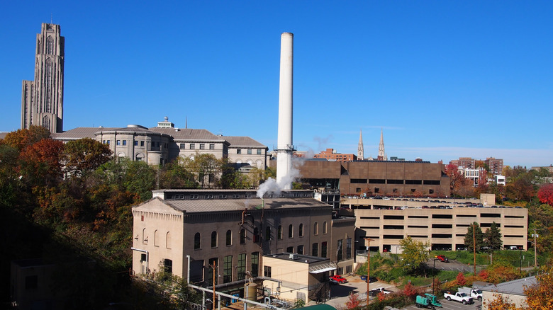 Oakland skyline, with the Cathedral of Learning on the left, the white stack of the Bellefield Boiler Plant in the center, and autumn trees around large white and brown buildings around them, with two distant church spires in the background, under a blue sky