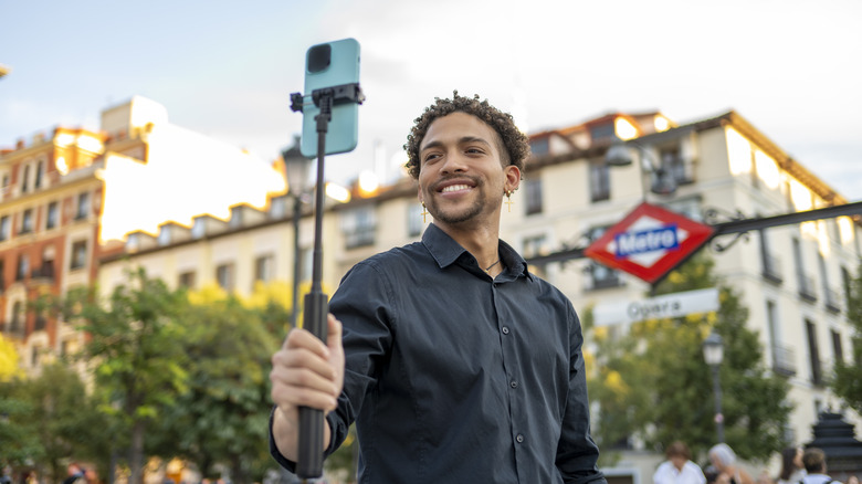 A man, smiling and holding a selfie stick, photographs himself in front of a metro station in Madrid, Spain