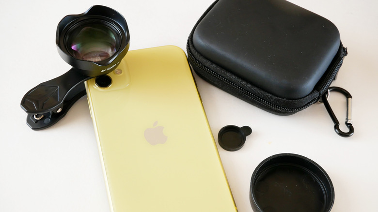 A close-up of a yellow iPhone with an external lens clipped over the camera and a black protective case beside it