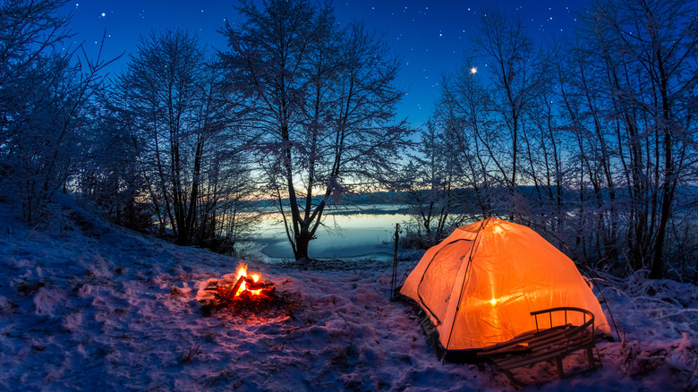 A glowing tent and campfire in a winter snowy scene with lake in background