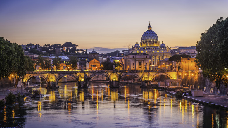 An ancient bridge over river and iconic Roman buildings at sunset