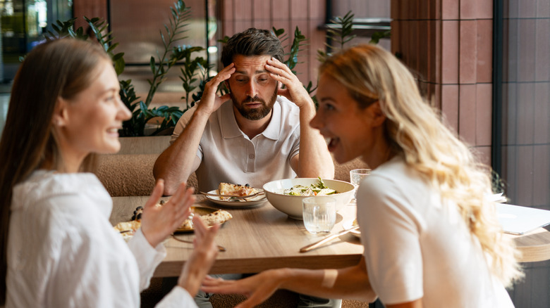 Two women talk loudly in front of a suffering man at a restaurant