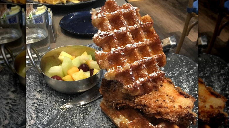A plate of chicken and waffles with a bowl of cut fruit