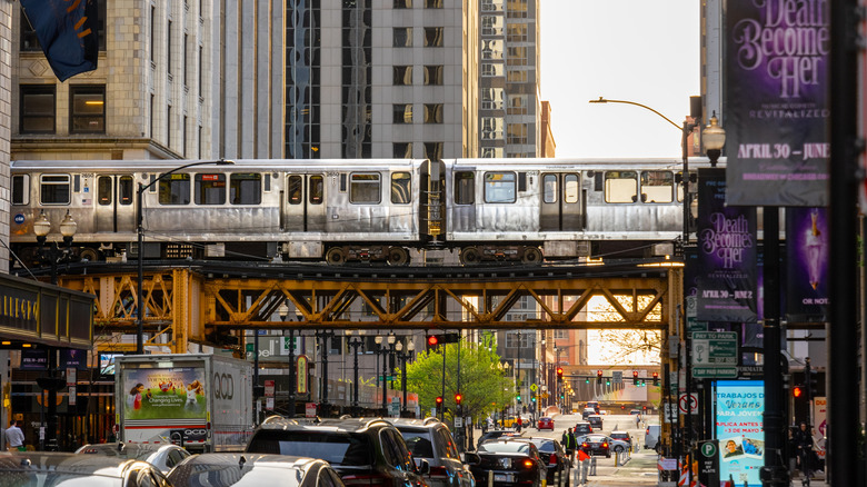 An elevated silver train crossing between buildings, with cars and tourists walking on the street below