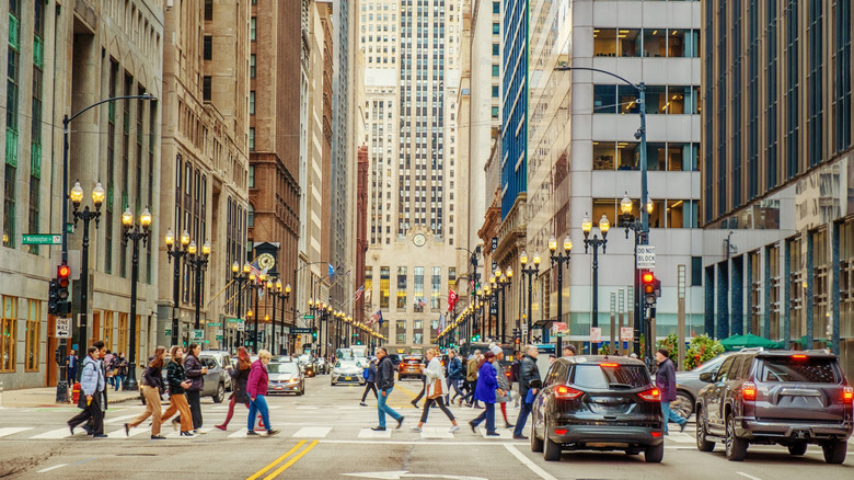 Cityscape of buildings on both sides and in the backdrop and people crossing a lamp-lined avenue