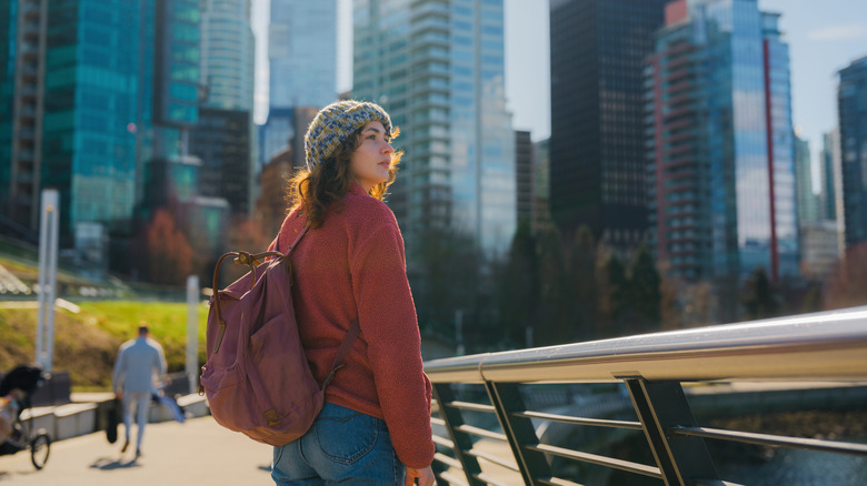 A girl with a beanie and backpack strolling along a waterfront with buildings in the backdrop