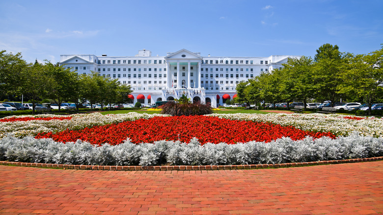 White front of the Greenbrier behind leafy trees and a display of red and white flowers, under a blue sky