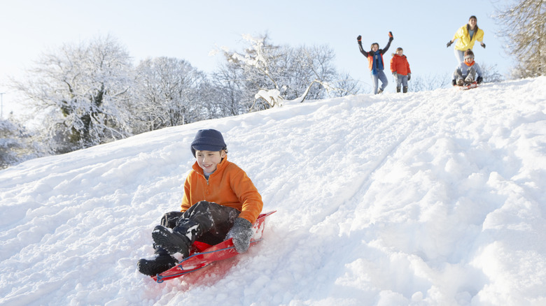 A young boy sledding down a snowy slope during winter with his family cheering behind him.