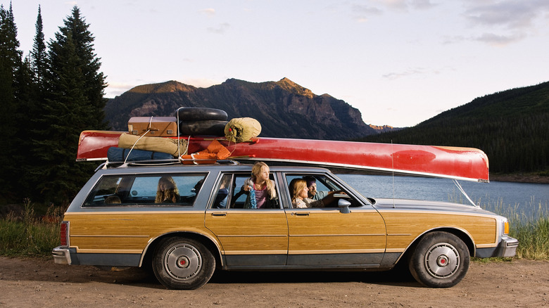 A family in a vintage station wagon with luggage and a canoe, with a lake and mountains in the background