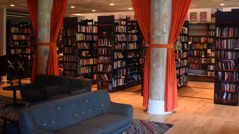 Alphabet City interior, with black shelves of books behind white columns with red curtains tied to them, and blue couches in the foreground on a colorful rug over a wooden floor