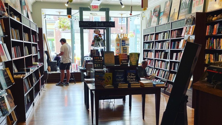 Inside Penguin Bookshop, with shelves of books along each wall and a table of books in the center of a wooden floor, one shopper crouched beside the shelf on the right, and another in a white shirt standing by the front window