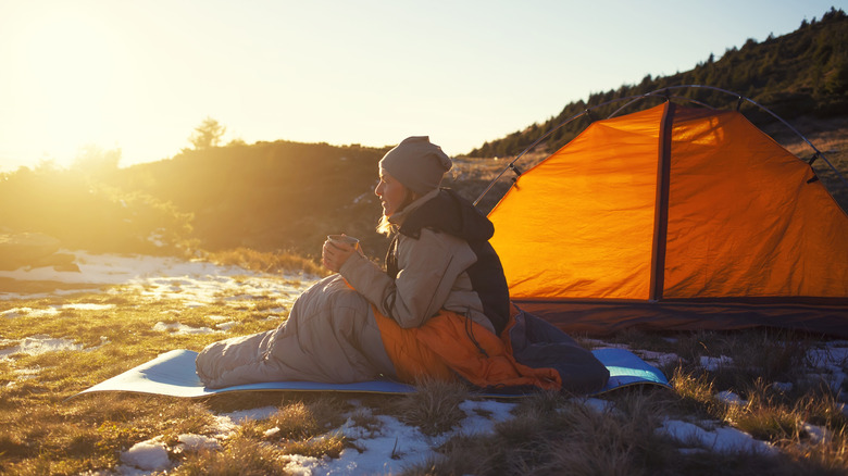 Woman sitting in a sleeping bag outside of a tent at sunrise, surrounded by melting snow