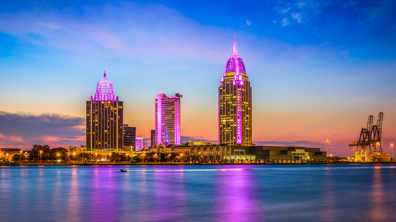 Large buildings in a skyline illuminated in purple that's reflecting onto water.