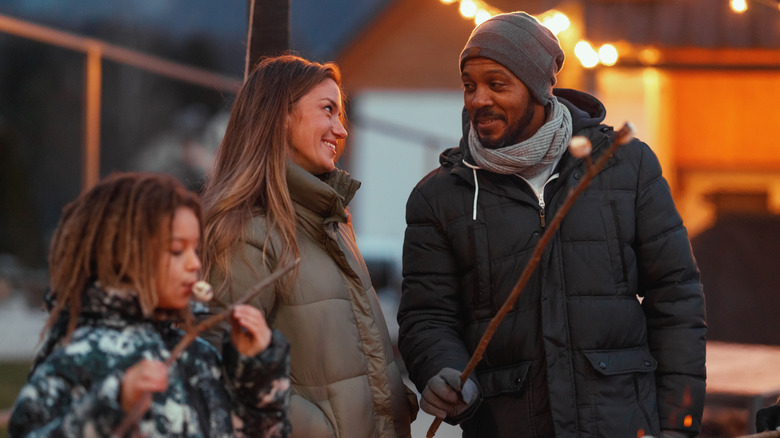 A happy family wearing winter jackets roasts marshmallows around a bonfire