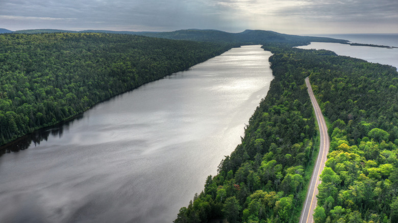 A small lake with a two-lane highway next to it surrounded by forest with Lake Superior on the horizon to the right