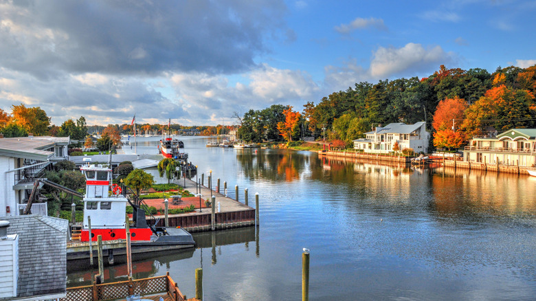 A river cuts through a colorful harbor, with the colors of fall spreading through the distant trees, and shiplap buildings and a red and white boat in the foreground.