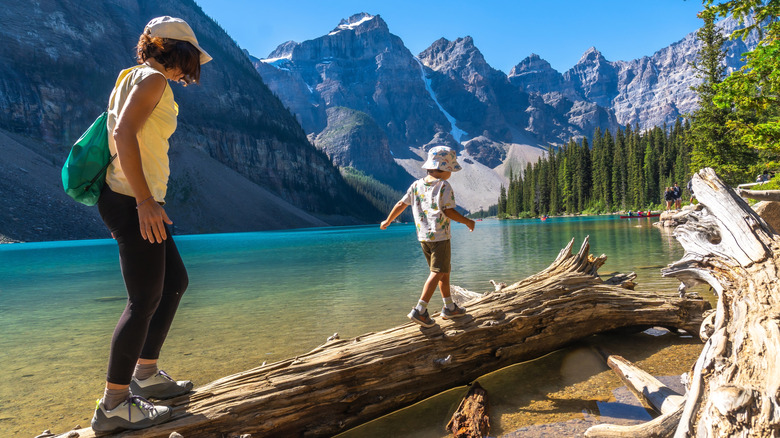 Woman and child cross fallen log with lake, mountains, and trees in background