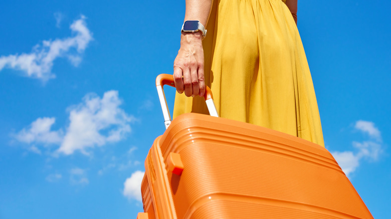 Woman in yellow dress holding orange suitcase handle against blue sky with sparse clouds