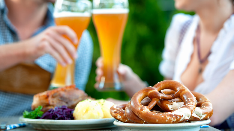 A couple clinking beer glasses with German food consisting of pretzels and pork knuckle are on the table