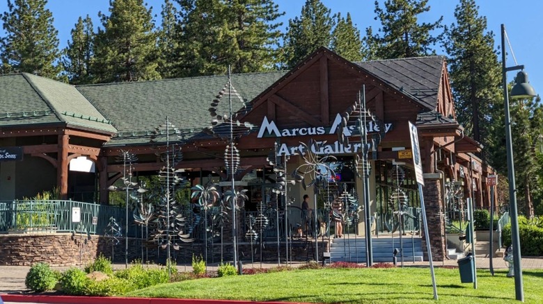 Iron wind sculptures fill the grass in front of dark-wood single story building with the sign Marcus Ashley Art Gallery in white lettering with pine trees and blue sky beyond.